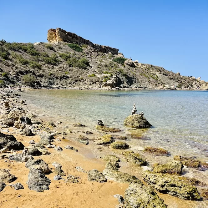 A rocky shoreline with scattered rocks and pebbles. The sea is calm, and there's a small stack of balanced rocks on a larger rock formation in the center. A rugged hill rises in the background under a clear blue sky.