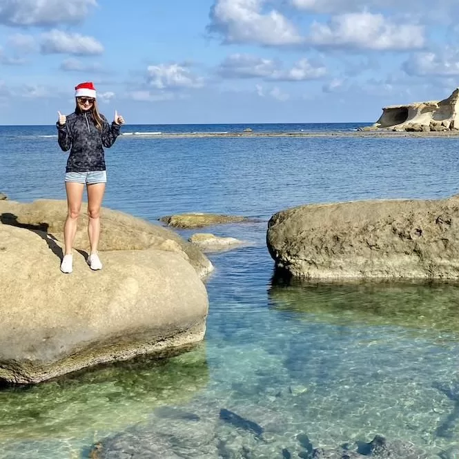 A person wearing a Santa hat and shorts standing on rocky terrain between clear, shallow waters. The person is smiling and giving thumbs up gestures. The sky is blue and sunny. 