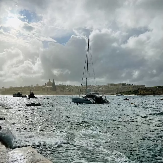 A sailboat anchored in a harbour with choppy waters. In the background, there are buildings along the coastline under a cloudy sky.