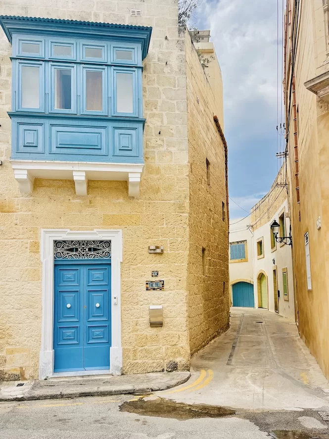 A narrow street in Naxxar adorned with traditional Maltese architecture, featuring a charming blue door and windows against a backdrop of limestone walls.