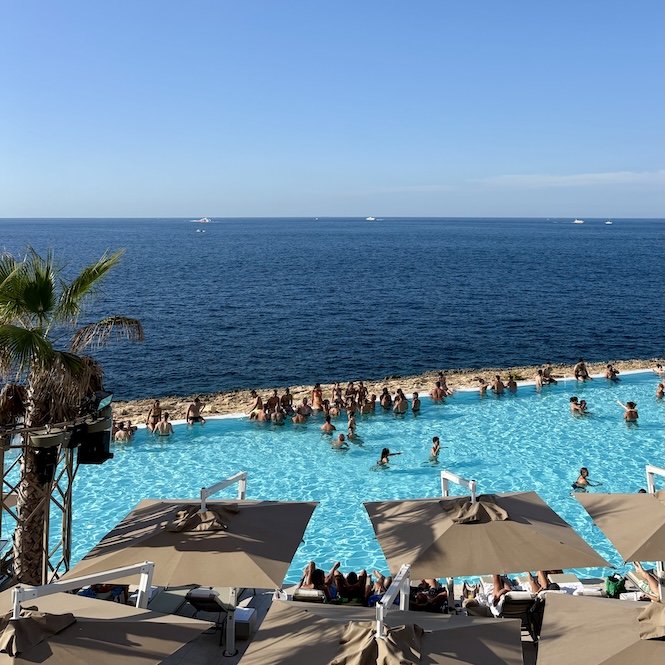 View of the infinity pool at Café del Mar in Bugibba, Malta, with people relaxing in the water and sunbeds under umbrellas, overlooking the open sea on a clear sunny day.