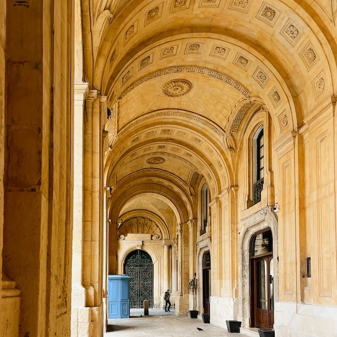 The grand arches and intricate ceiling details of the National Library of Malta, Bibliotheca Nazionale, are highlighted in this image from a Valletta walking tour. The warm sandstone corridors and the classic blue wooden door at the end create a path leading to historical and literary discovery.