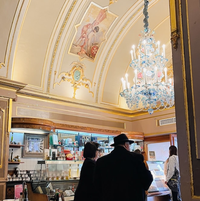 Visitors inside the historic Caffe Cordina in Valletta, captured during a walking tour. The interior features ornate golden ceilings with fresco paintings, an exquisite blue and white chandelier, and a traditional Maltese café counter.