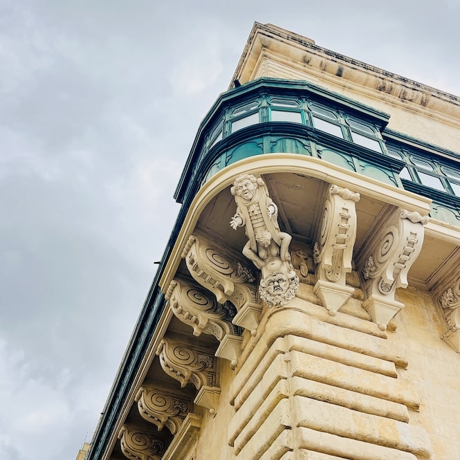 An architectural detail from a Valletta walking tour, showing a grand limestone balcony with ornate corbels and a unique sculpture of a clown on a naked woman, symbolizing the Grandmaster's defiance. This historical anecdote adds a layer of intrigue to the ornamental façades of Valletta.