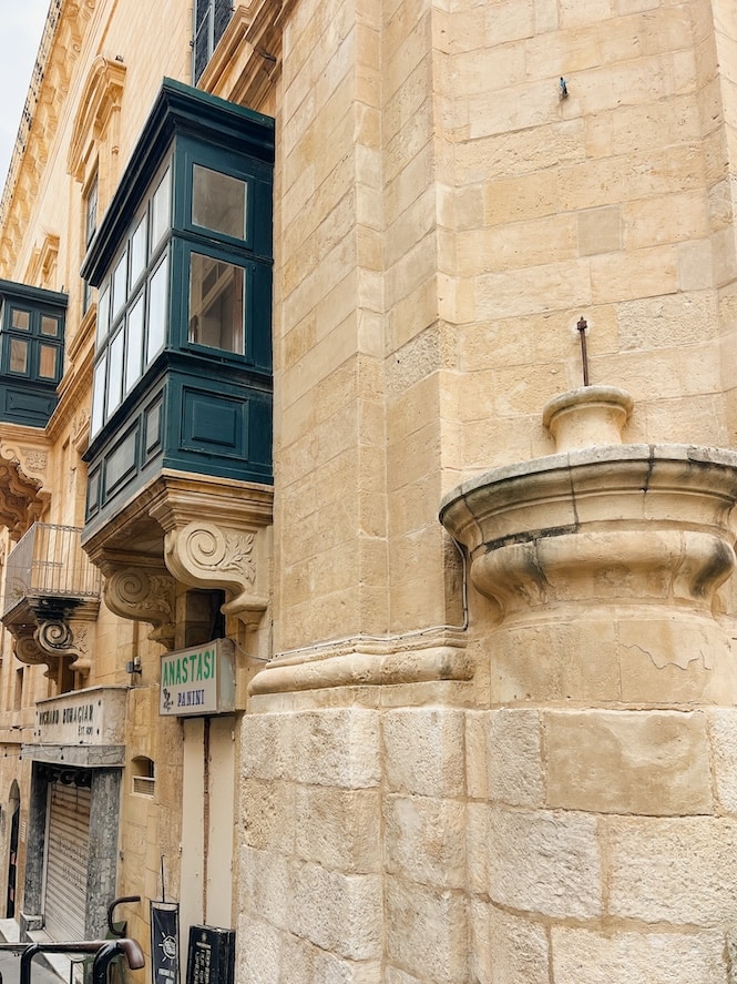 GPT A traditional Maltese balcony extends from a weathered limestone building at a notable shaming corner on the Valletta Walking Tours route, Malta.