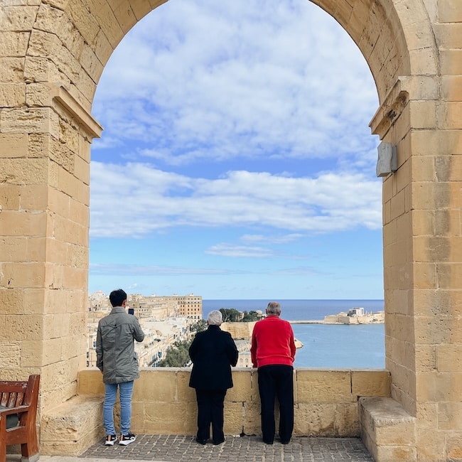 Three people gaze out at the panoramic view of the Mediterranean Sea from the Upper Barrakka Gardens in Valletta, during a Valletta walking tour. The large stone archway frames the serene blue waters and the historic cityscape, inviting a moment of reflection and appreciation for Valletta's beauty.