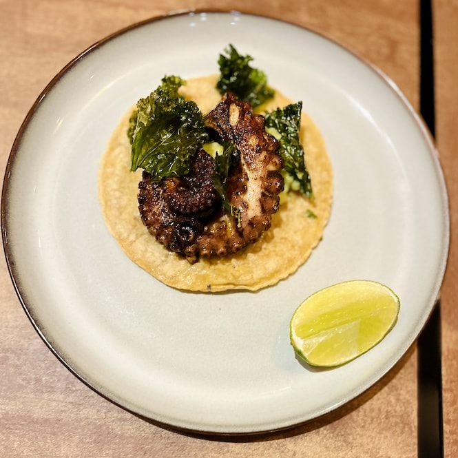 A plate with octopus tacos served on a soft corn tortilla, garnished with crispy kale and accompanied by a lime wedge.