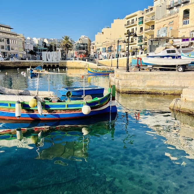 Luzzu boats docked in Spinola Bay, with colorful buildings and boats reflected in the calm, turquoise water.