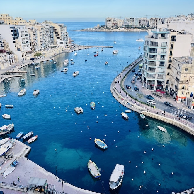 Aerial view of Spinola Bay in St. Julians, Malta, with boats floating on clear blue waters surrounded by buildings and cars along the waterfront.
