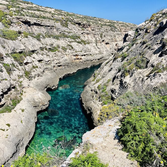 The canyon is in the Ghasri Valley in Gozo, surrounded by cliffs and greenery.