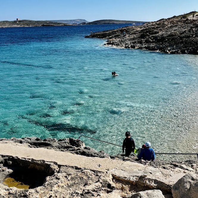 Ħondoq Bay overlooks the island of Comino. Two people sit on the rocky coast and enjoy the bright blue sea.