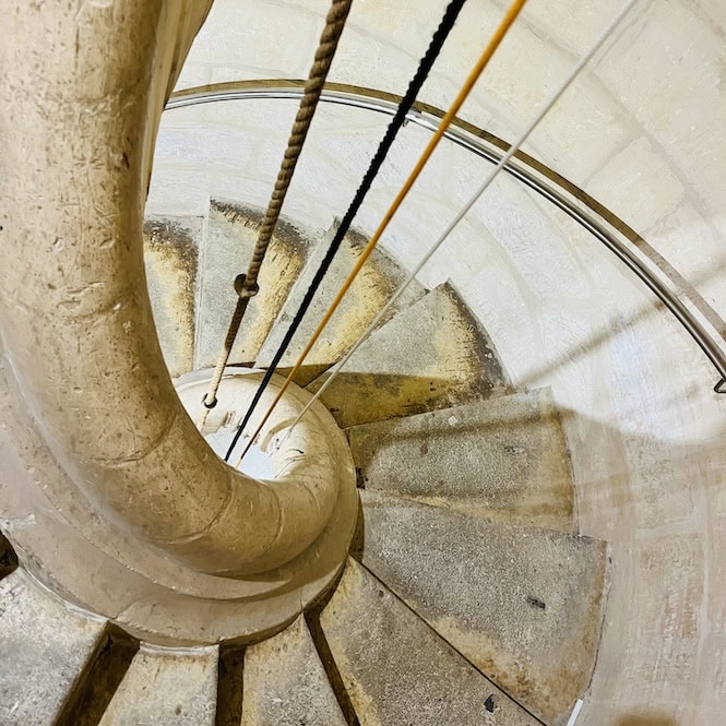 A close-up view of the ancient spiral limestone staircase inside the Mosta Dome, with well-worn steps curving upwards towards the balcony, illuminated by soft natural light streaming through the stone walls.