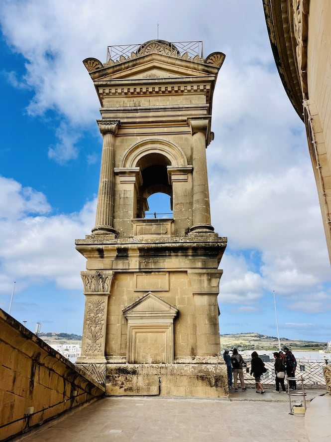 A view of the ornate Mosta Rotunda balcony, with its intricate stone carvings and columns, offering panoramic views of the surrounding landscape and cityscape.