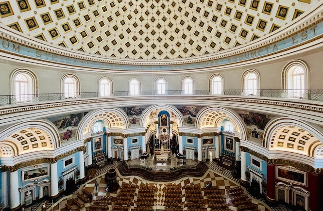 Interior view of the Mosta Rotunda church in Malta, showcasing the grand circular nave with an ornate domed ceiling featuring intricate geometric patterns. The church is adorned with multiple balconies, arched windows, and a beautifully decorated altar area at the far end.