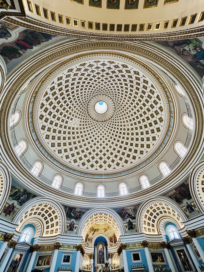 A stunning interior view of the Mosta Dome, showcasing the intricate geometric patterns and ornate details of the massive unsupported dome, with natural light illuminating the grand space through windows along the base of the dome.