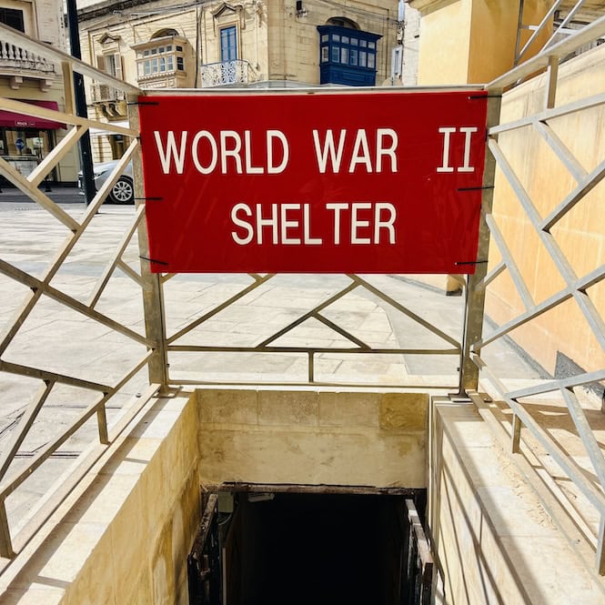 A sign reading "WORLD WAR II SHELTER" marks the entrance to underground bomb shelters from the Second World War, located outside the Mosta Rotunda church in Malta. The entrance is framed by a metal railing leading down concrete steps.
