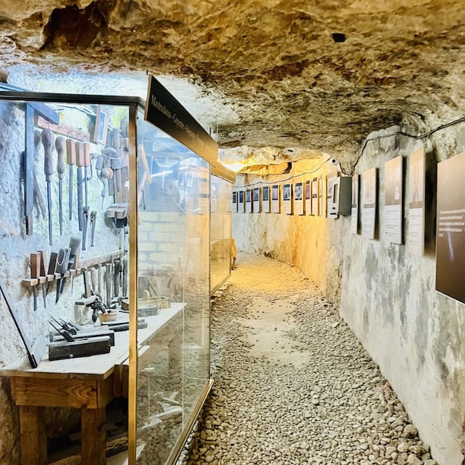 The interior of the World War II air raid shelters outside Mosta Rotunda in Malta, showing a narrow passageway lined with stone walls and display cases containing artifacts. The rough, rocky ceiling and floor are visible, illuminated by artificial lighting.