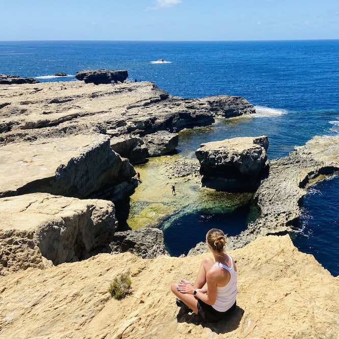 I see a person sitting on the rocky coastline overlooking the Blue Hole. The rugged coastline features layered, flat rock formations that jut out into the deep blue Mediterranean Sea.