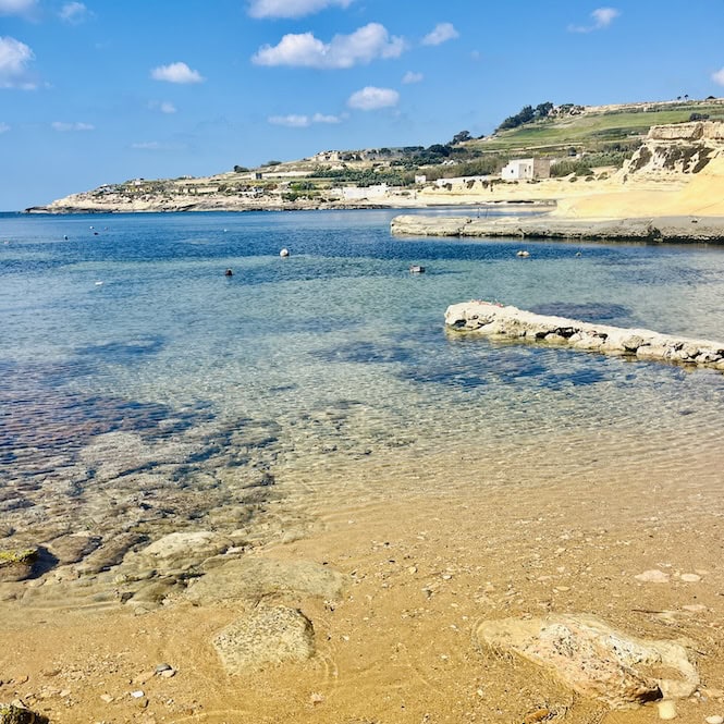 Crystal clear turquoise waters of Xatt l-Aħmar Bay on the island of Gozo, with rocky shore and hills in the background.