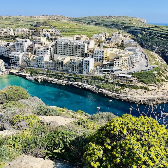 Aerial view of the picturesque coastal village of Xlendi on the island of Gozo, with its vibrant blue bay, rocky cliffs, and charming white buildings, as seen from a hiking trail.