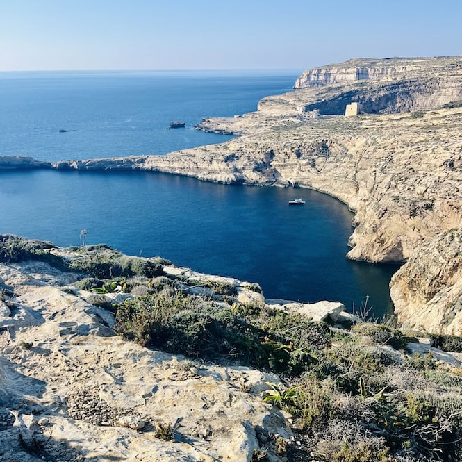 Spectacular view of Dwejra Bay on the island of Gozo, featuring rugged limestone cliffs, crystal-clear turquoise waters, and small boats, as seen from a scenic hiking trail.