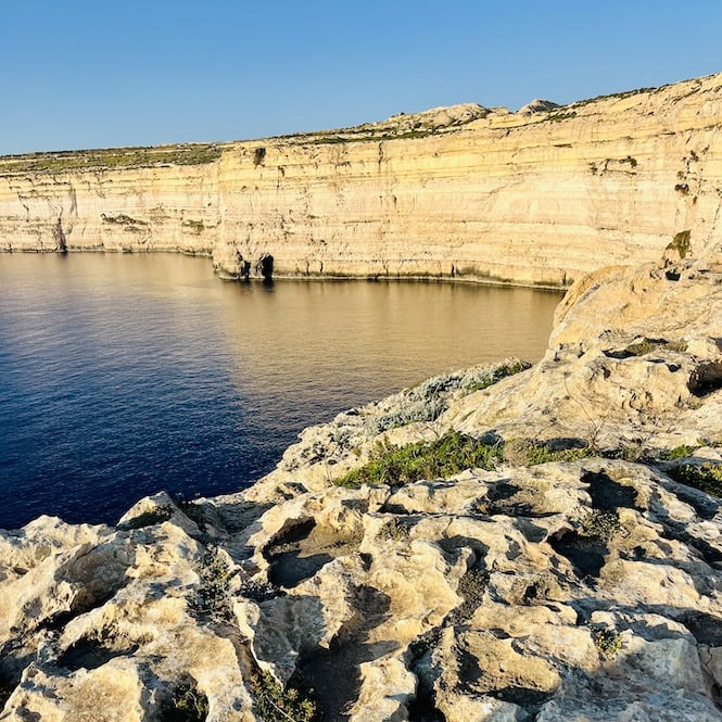 Rocky coastline with tall cliffs on Gozo's west overlooking calm blue sea.