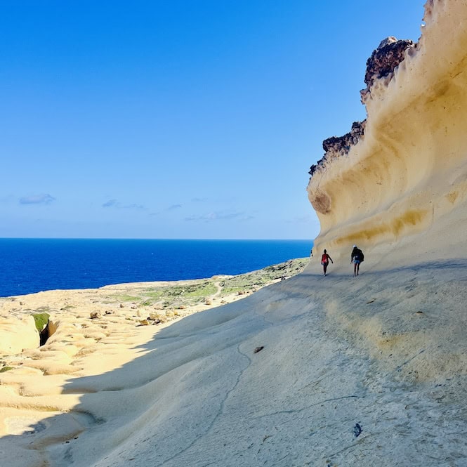 Hikers exploring the dramatic sandy coastline at the base of tall limestone cliffs on the island of Gozo, Malta.
