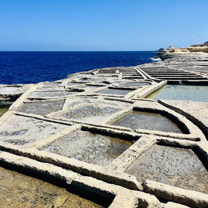 Ancient salt pans carved into limestone rock overlooking the deep blue Mediterranean Sea under a clear sky on the island of Gozo, Malta.