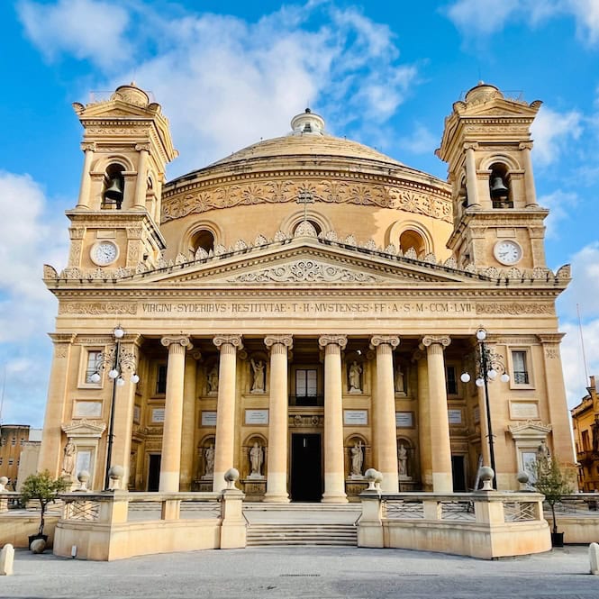 Exterior view of the majestic Mosta Dome, also known as the Rotunda of Mosta, showcasing its ornate Neoclassical architecture and grand facade against a bright blue sky with wispy clouds.
