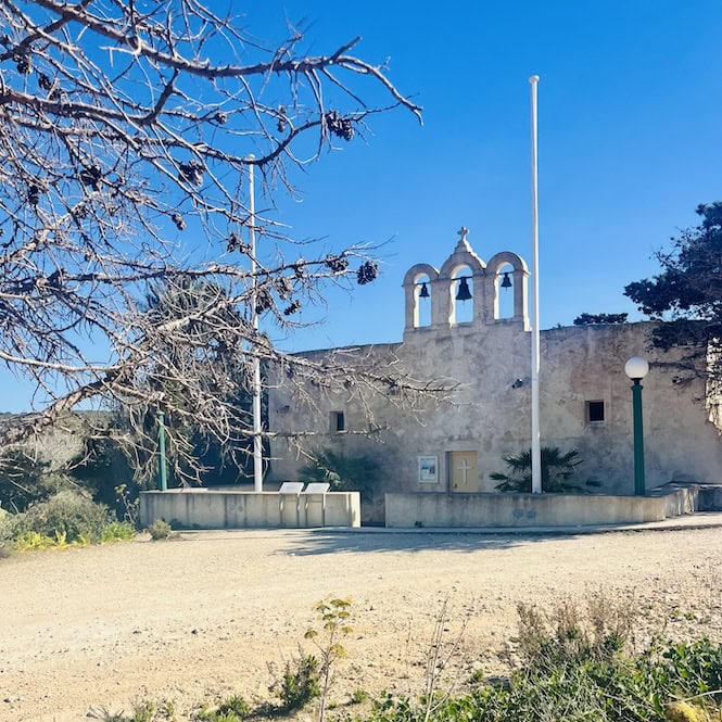 An old limestone church building with bell towers stands amid bare trees on a sunny day, along the Comino coastal hike trail.