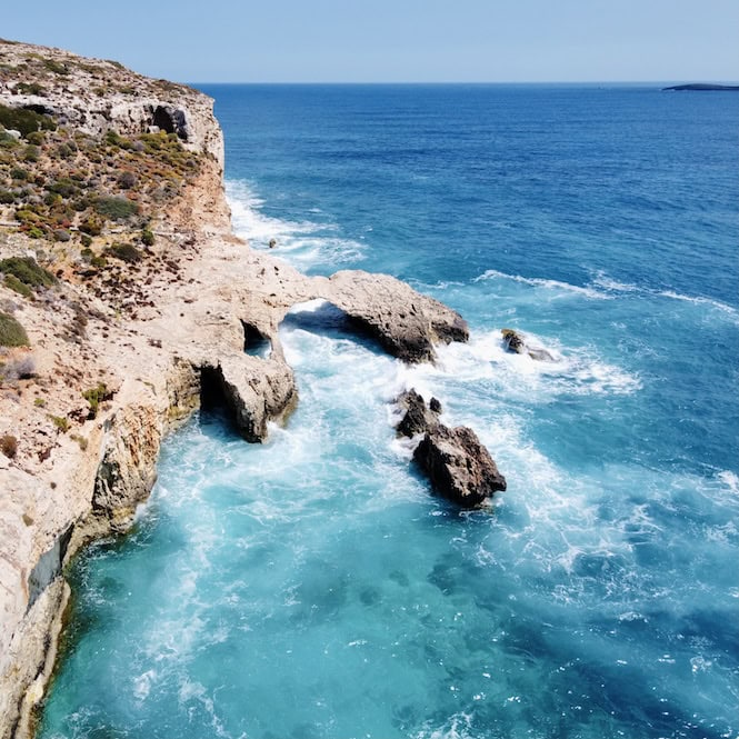 Turquoise waters crash against the rugged coastline of Comino, Malta, revealing sea caves carved into the cliffs.