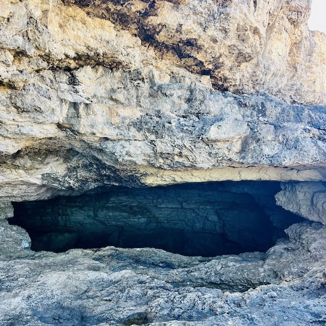 The entrance to a dark cave with layered rock walls, seen while hiking the coastal trail on Comino island.