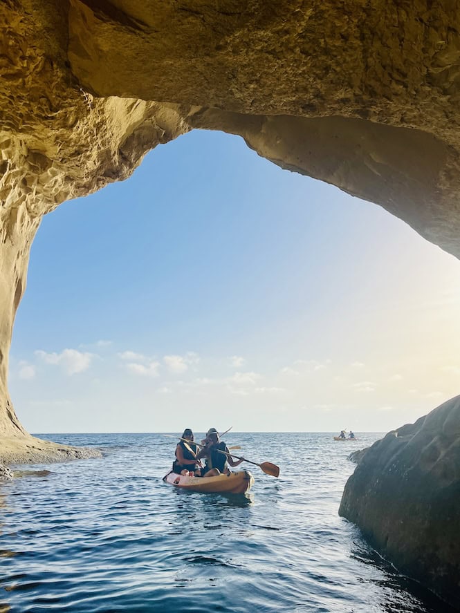 A group of people kayak through the waters near Ta' Marija Cave at Gnejna Beach in Malta, with sunlight illuminating the cave's archway and the serene sea beyond.
