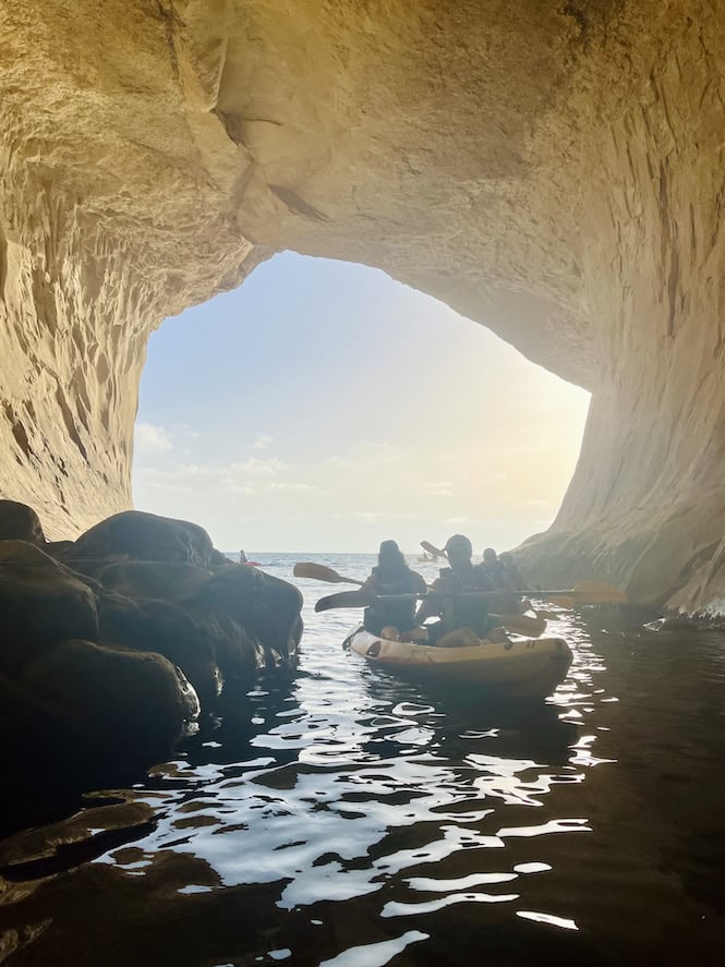 Kayakers paddle through the tranquil waters inside Ta' Marija Cave near Gnejna Beach, Malta, framed by the cave's large archway with sunlight glinting off the water and highlighting the natural rock formations.