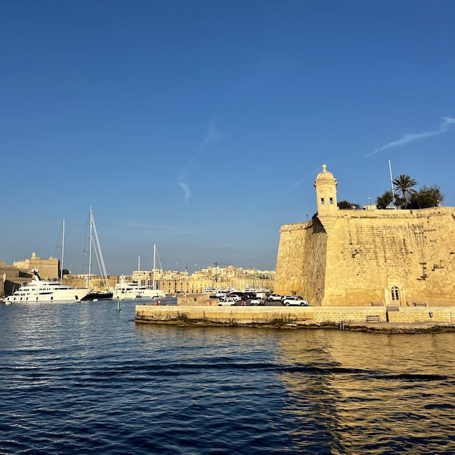 Boats docked in front of a historic limestone fortification with a watchtower in Gardjola Gardens in Senglea, part of Malta's Three Cities.