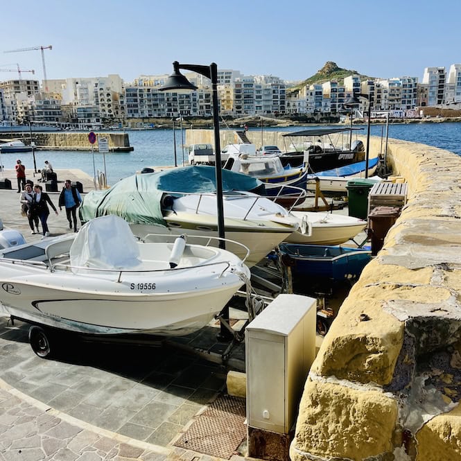 Several small boats docked along the waterfront in the harbour of Marsalforn, Gozo, with residential buildings visible in the background.