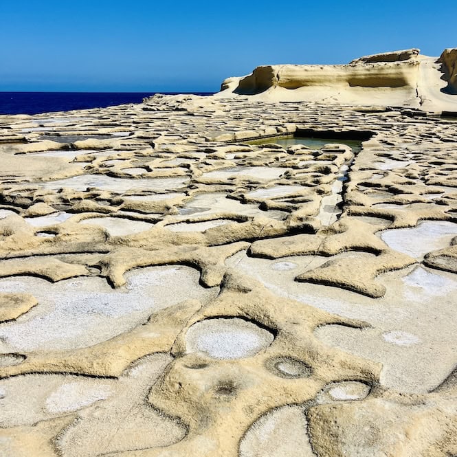An eroded and pitted rocky shoreline in Marsalforn, Gozo, with small pools of seawater settled in the salt pans, and the blue sea visible in the background.