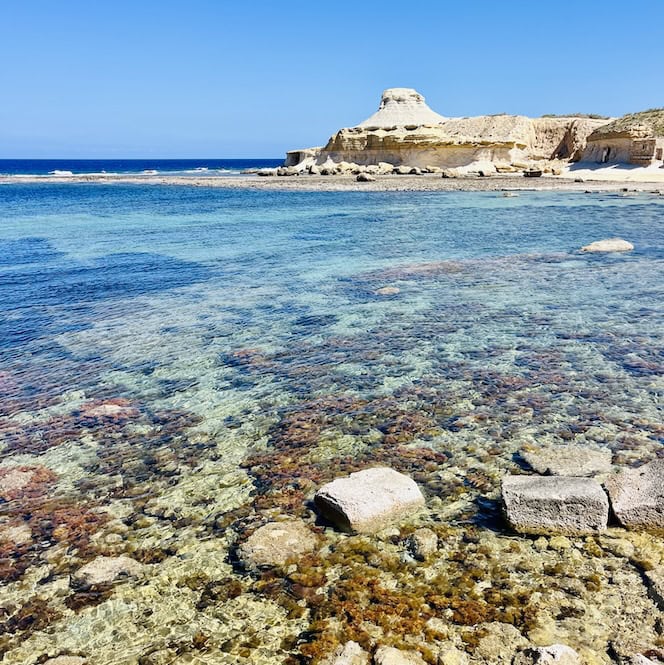 Clear turquoise waters at Xwejni Bay, close to Marsalforn, Gozo, with the rocky shoreline and cliffs visible in the distance under a bright blue sky.