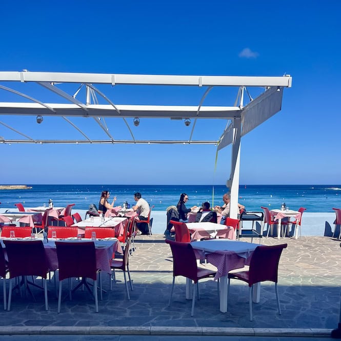 People eating and socializing at red tables under a white canopy structure at an outdoor dining area overlooking the blue sea in Marsalforn, Gozo