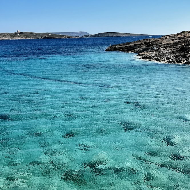 Clear shallow waters over sandy seabed, with a watchtower on the rocky coastline of Comino in the distance.