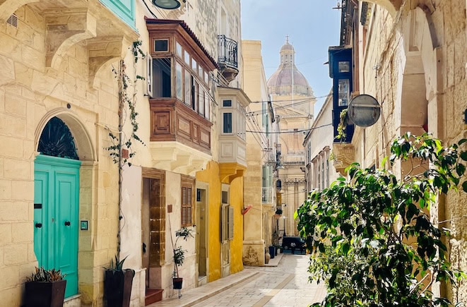 A narrow, colourful street in Birgu, one of Malta's Three Cities, with traditional Maltese houses, wooden balconies, and a dome of a church visible in the background. This image is captured on a Three Cities walking tour.