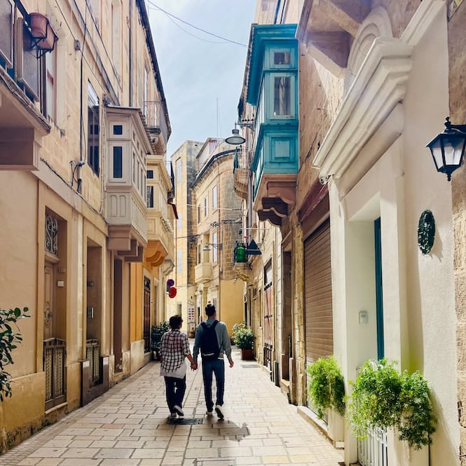 Two people walking down a picturesque, narrow street in one of Malta's Three Cities, surrounded by traditional Maltese architecture with colorful doors, windows, and balconies.