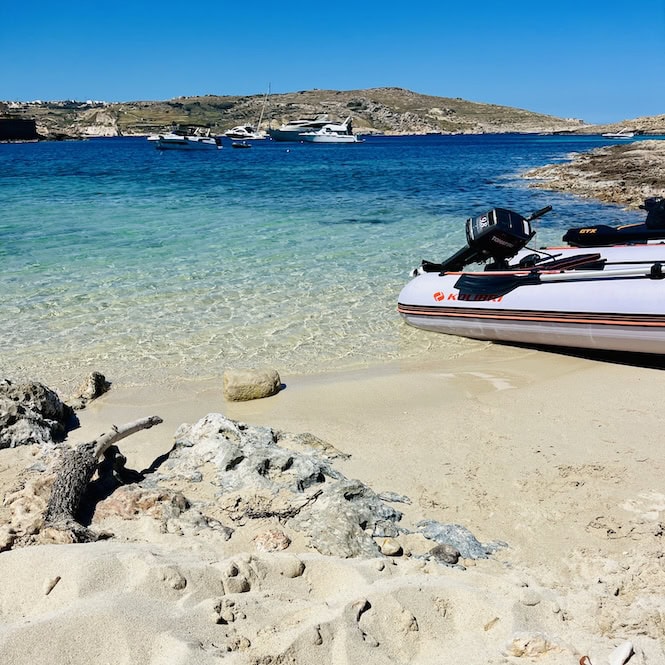 A boat is docked in the clear turquoise waters of the Santa Maria Bay on Comino, with rocky cliffs and beach in the foreground, a popular stop on the island's coastal hike.