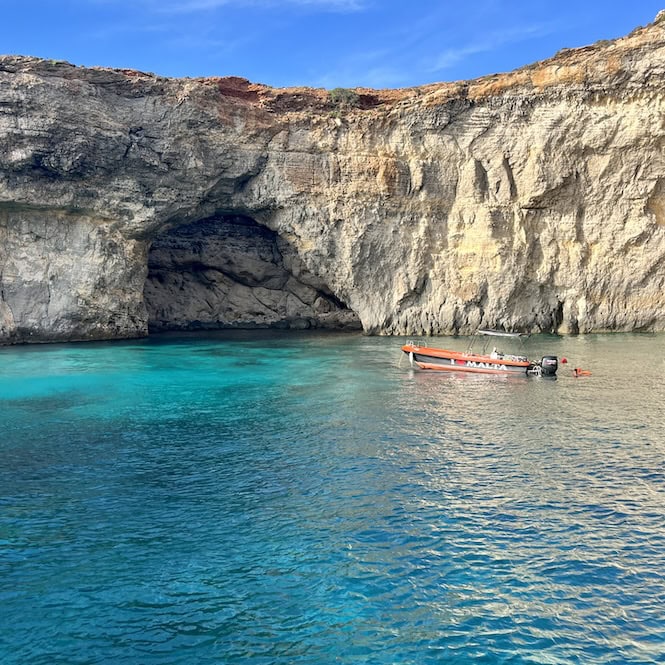 A small boat floating in the turquoise waters by the towering cliff caves in Malta, capturing the island's natural beauty in September.