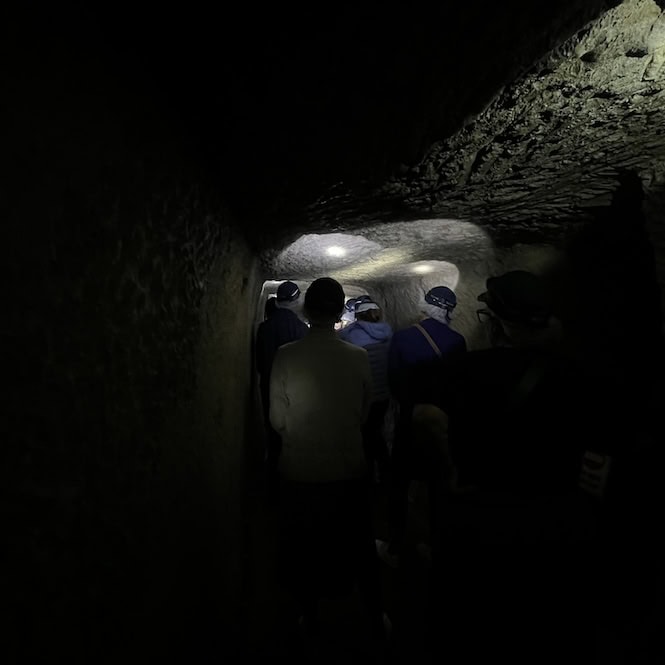 A group of people with helmets are listening to a guide. They are in dim light in a dark tunnel.