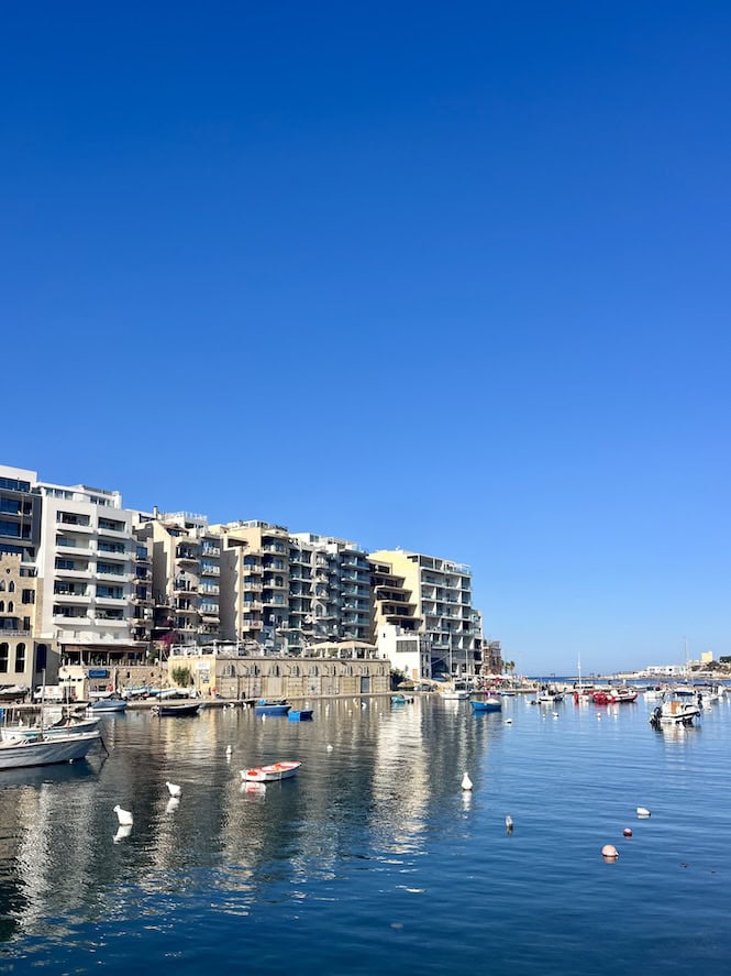 A picturesque view of Spinola Bay in St. Julian's, Malta, featuring modern apartment buildings along the waterfront. Boats are docked in the calm bay under a clear, vibrant blue sky. St. Julian's is known as one of the best areas to stay in Malta for nightlife and restaurants.