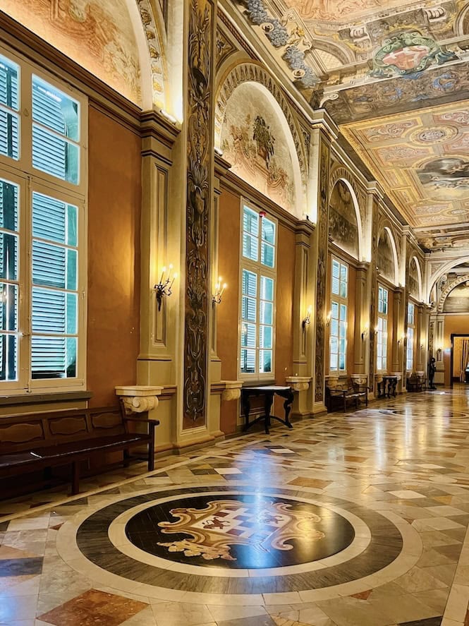 An opulent interior hallway with intricate ceiling frescoes, large arched windows, marble floors with an inlaid design, and elegant wooden doors in the Grandmaster's Palace in Valletta, Malta.