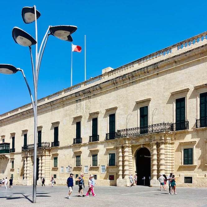 The exterior facade of the Grandmaster's Palace, a major landmark in Valletta, Malta, showing ornate balconies, columns and arched windows. Modern street lamps are in the foreground and people are walking through the square in front of the palace.