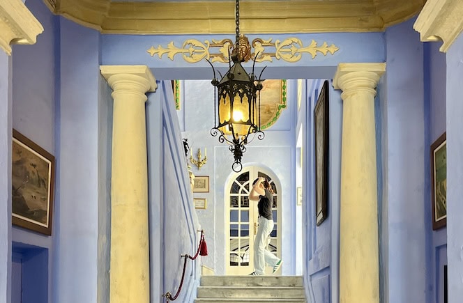 This image captures the interior of Casa Rocca Piccola in Valletta, Malta. The vibrant blue walls and grand columns frame a staircase adorned with antique paintings and a traditional hanging lantern, showcasing the historic and elegant design of this 16th-century palace.