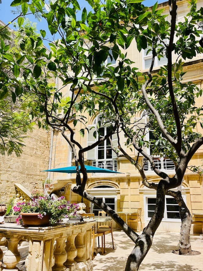 This image shows the lush garden of Casa Rocca Piccola in Valletta, Malta. The courtyard features vibrant green trees, blooming flowers, and a shaded seating area, all set against the backdrop of the historic limestone facade of the palace.